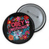 A black round pinback button featuring floral embroidery-style designs and bold red text that reads "Obey Nothing."